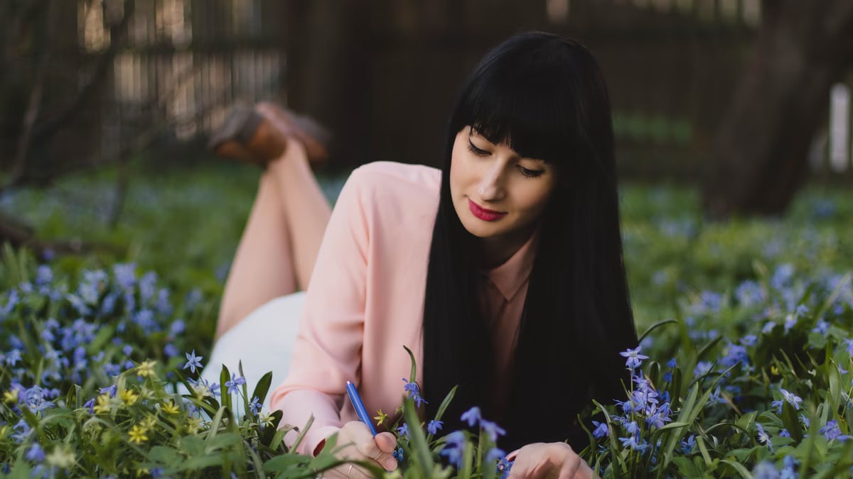 A woman writing in a journal while lying in a field of purple and yellow flowers outdoors.